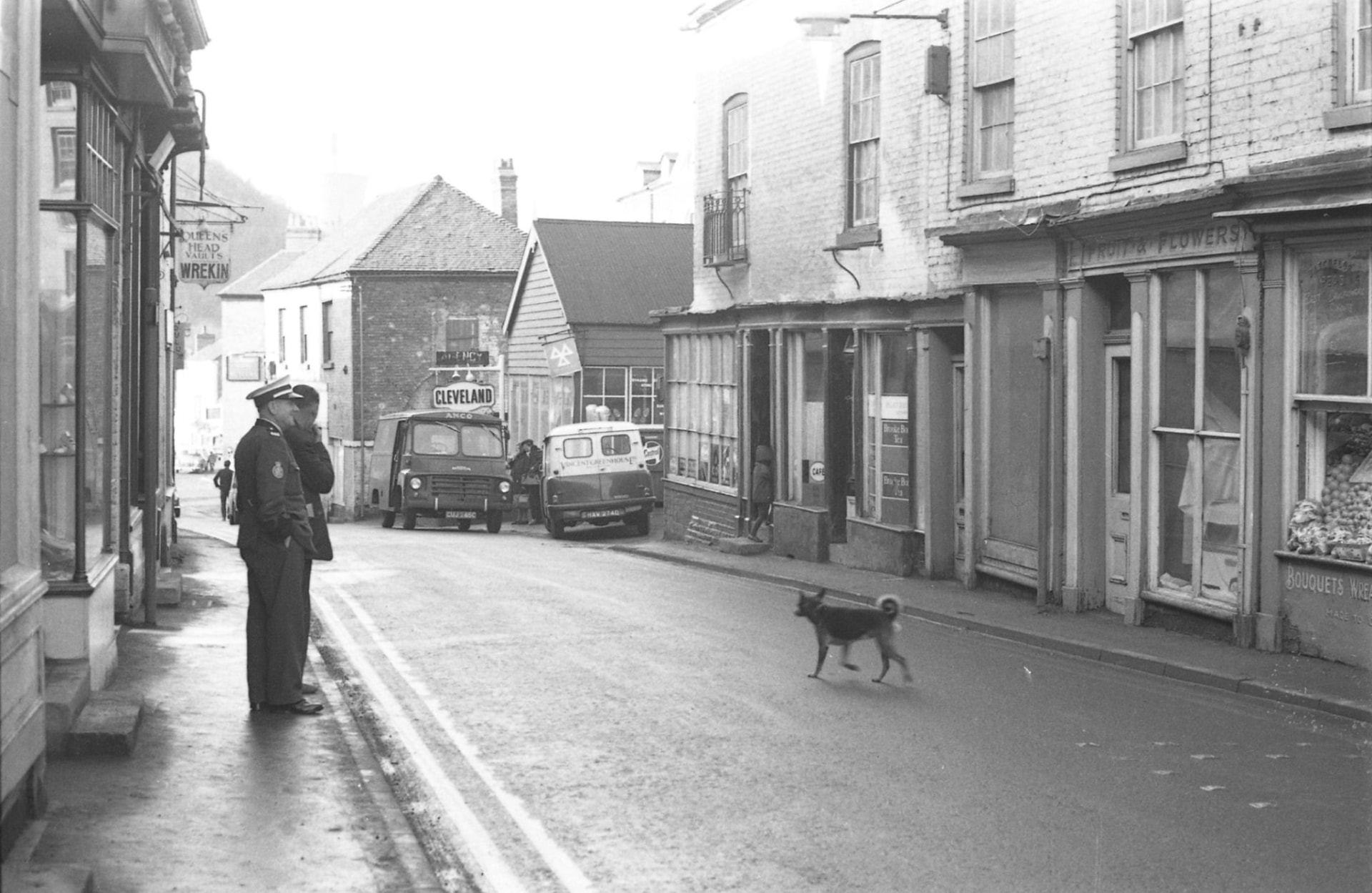 High Street, Ironbridge: the Queen's Head Vaults Wrekin sign hanging on the left, with shops, delivery vans and a dog crossing the road.