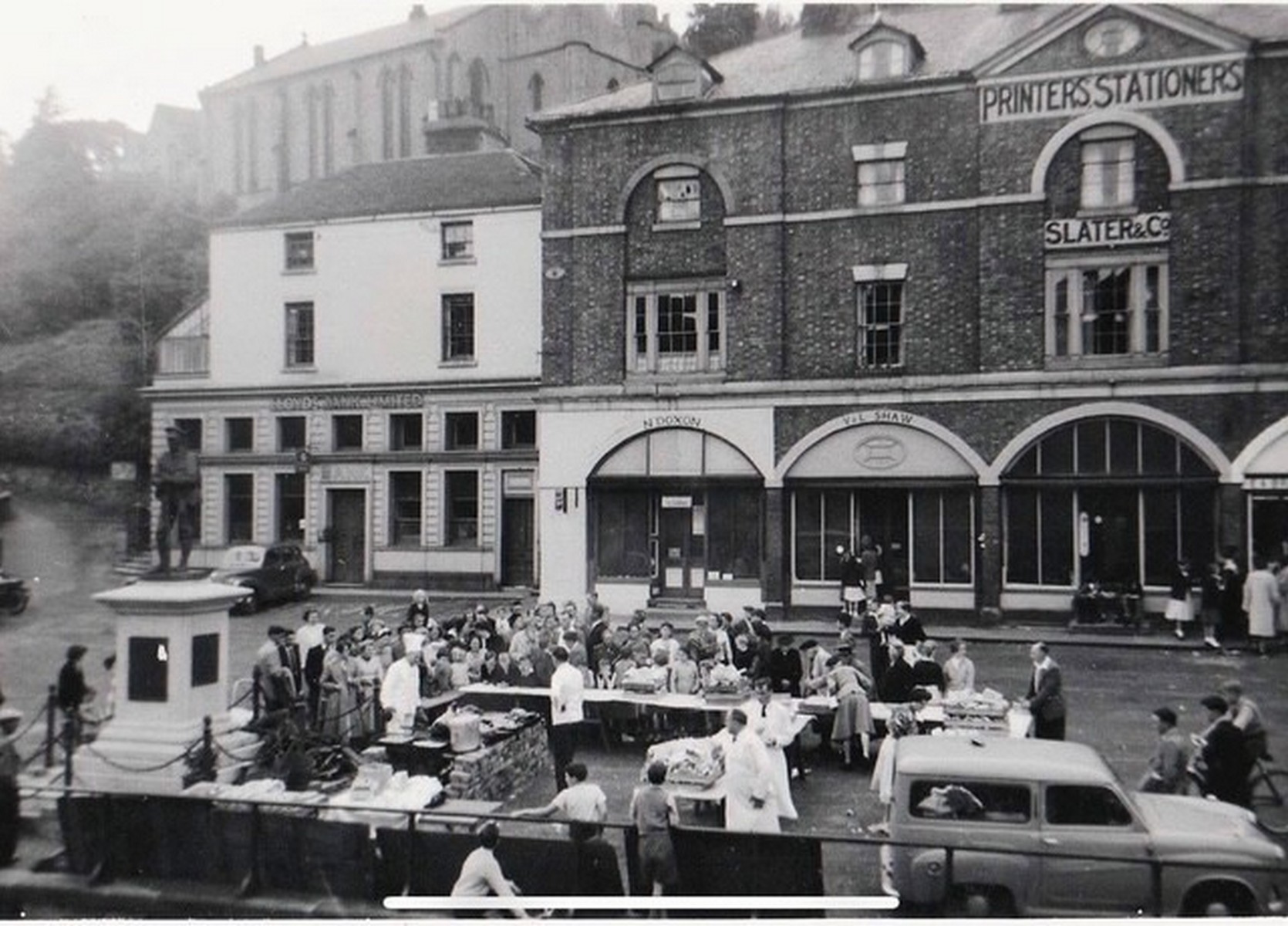 Market day below the same frontage, N Doxon and V & E Shaw signed over two of the arches and Lloyds Bank standing to the left. Trade spilled straight out of the arches onto the stalls in front — shop and market were one business, not two. Exact date unknown.