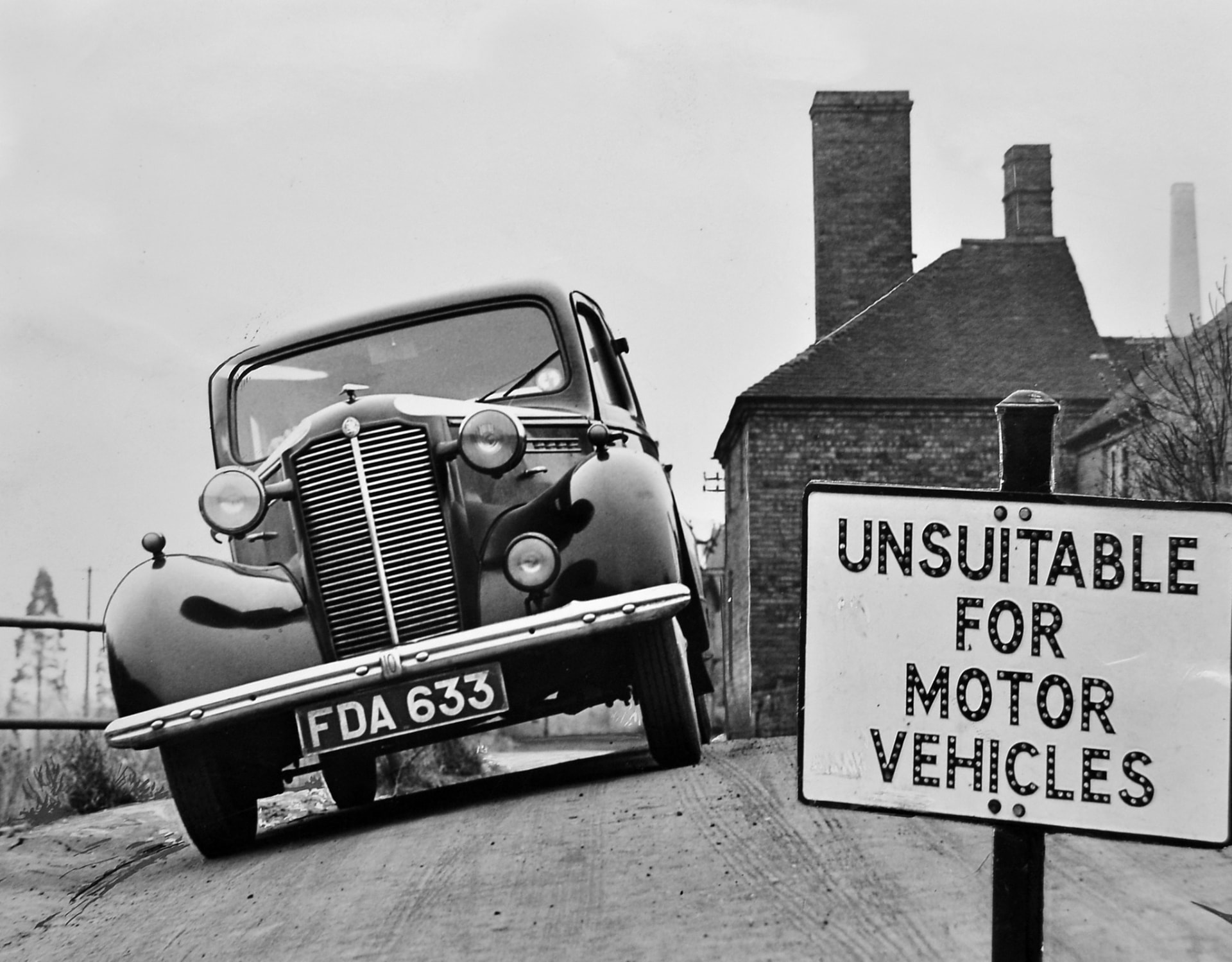 A car leans beside an 'Unsuitable for Motor Vehicles' sign as the road surface gives way.