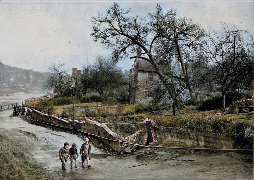 Three boys walk the buckled riverside road past a collapsed retaining wall. Later colourised.