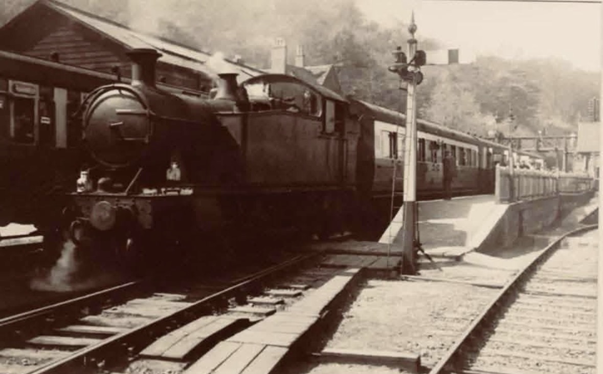A tank engine waits alongside the platform with its carriages in this early 20th-century view, the down starter signal standing sentinel beside the track. It's one of the clearest surviving images of platform edge, signal and station awning together, capturing the everyday rhythm of a working country station before living memory.