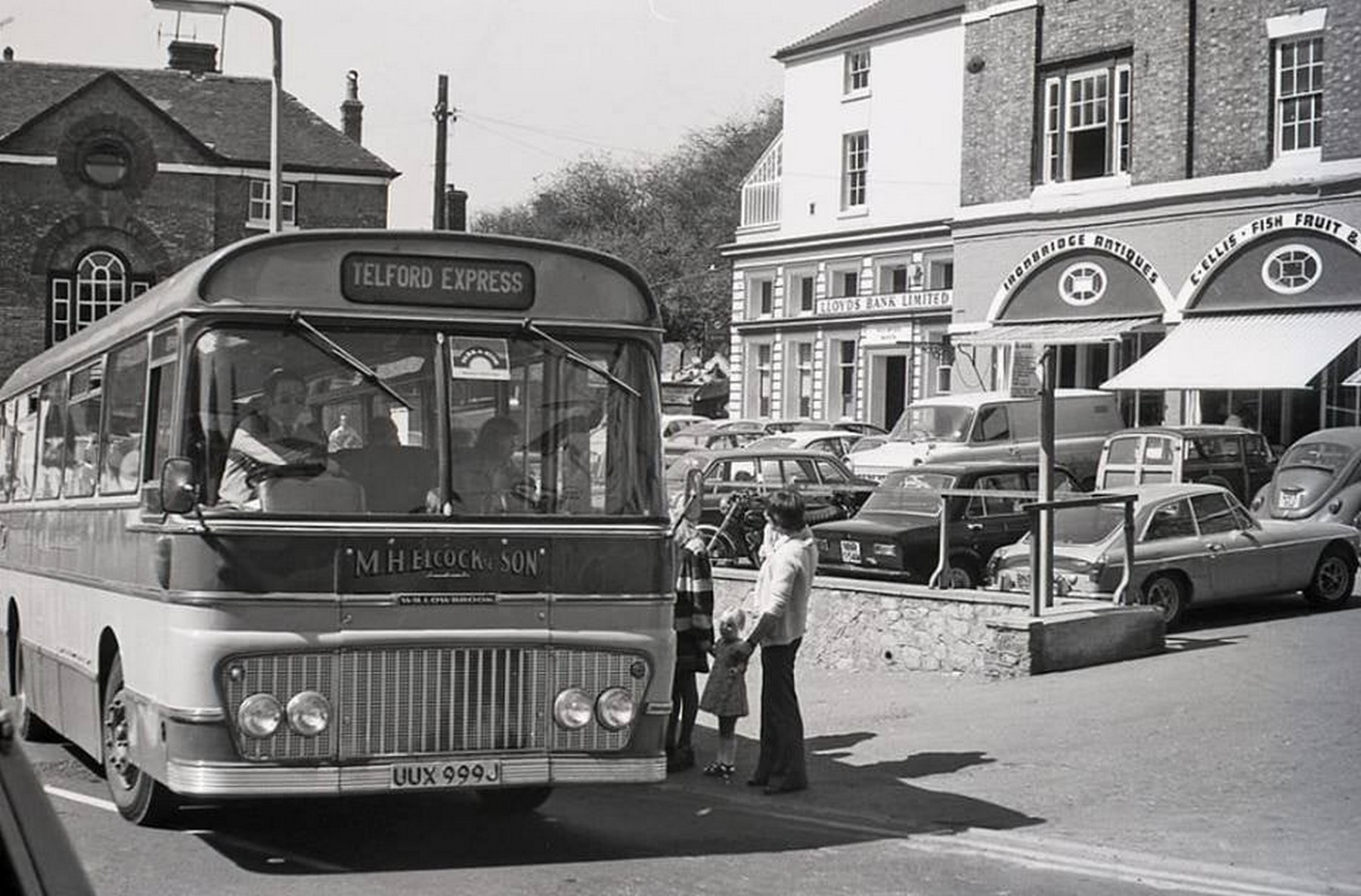A Telford Express coach at M H Elcock & Son, with Lloyds Bank and Ironbridge Antiques behind. The antiques shop is the telling detail: by the early 1970s the Gorge was beginning to sell its own past to visitors. Early 1970s.