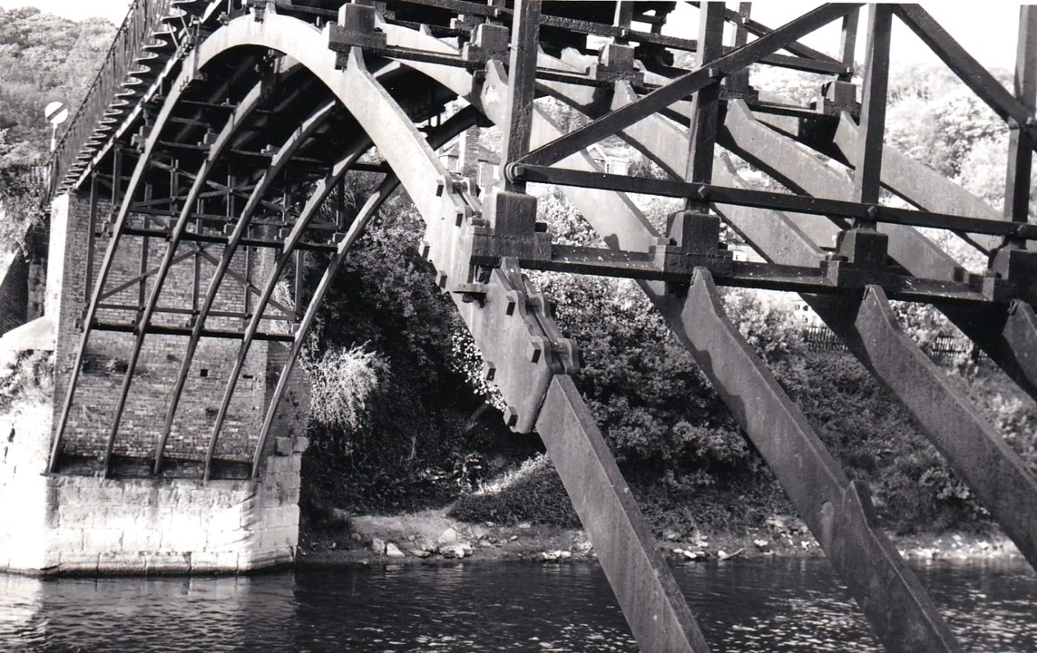 Photographed from the water's edge sometime in the twentieth century, this study captures the cast-iron arch ribs and their radiating spandrel bracing in close detail. Look for the slender paired ribs carrying the deck — John Onions' 1818 design cleverly reused the older abutments beneath, saving cost while giving the bridge its distinctive lightness.