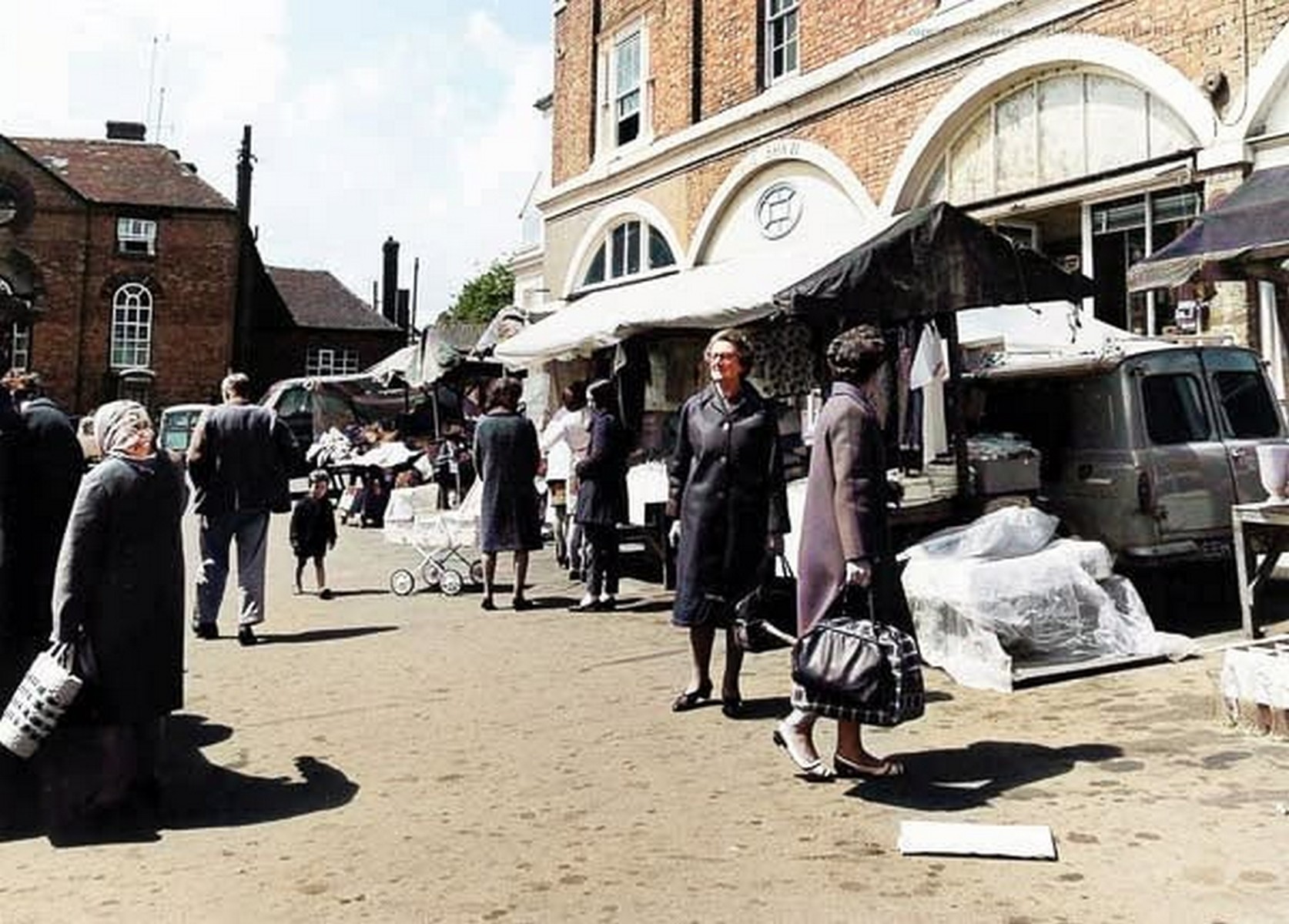 A busy market morning with awnings run out along the whole length of the Square. Follow the line of canvas and you can see just how far the stalls once reached — right across ground now given over to parking. Colourised; exact date unknown.