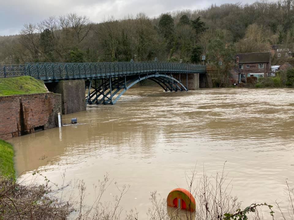 The Severn in flood, brown water pushing hard against the bridge's abutments in this modern photograph. Floods are nothing new to this site — one destroyed the middle pier of the earlier timber bridge back in 1795 — and the river continues to test the iron structure's foundations to this day.