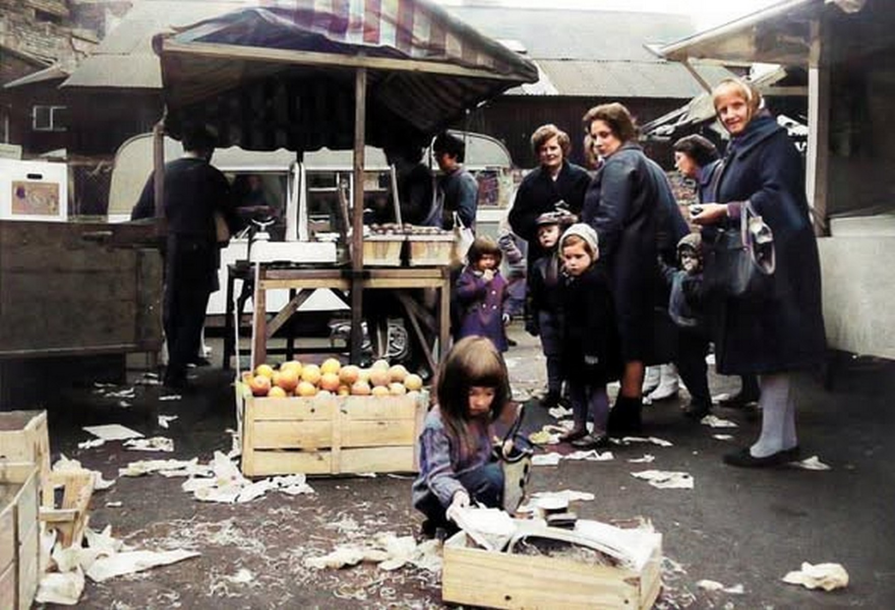Shoppers gathered round the fruit and vegetable crates, headscarves knotted and winter coats buttoned. The produce is still in its wooden transit crates — no packaging, no refrigeration, just what had come in that morning. Exact date unknown, but the dress places it in the post-war years.