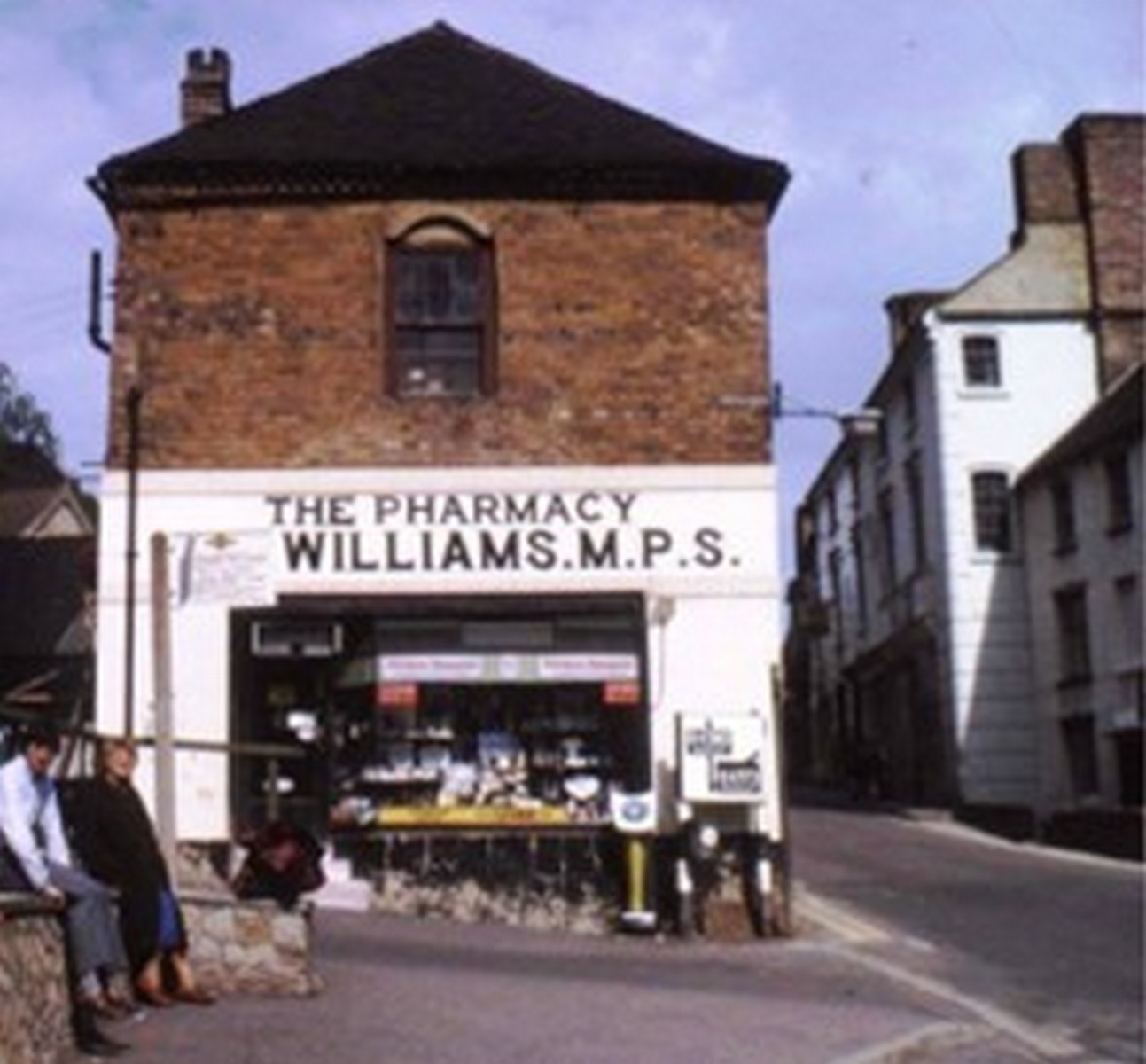 Williams the chemist, lettered "THE PHARMACY — WILLIAMS M.P.S." across the fascia. This is the same Market Hall building that houses the pharmacy today, seen at an earlier stage with a different shopfront and painted sign. Note the low stone wall at the left, still there now. Exact date unknown.