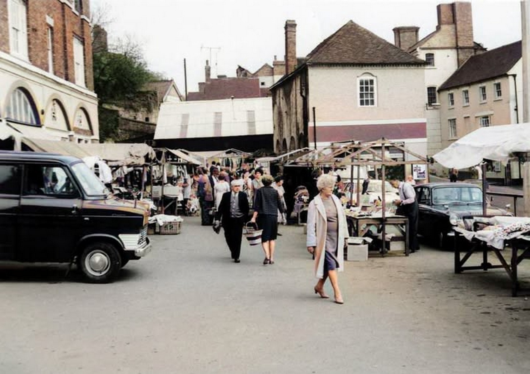 Market day in the 1970s, a delivery van pulled up tight against the stalls. Goods now arrived by road rather than by cart or river, and the van doors double as the back of the stall — a practical fix traders across Shropshire adopted.