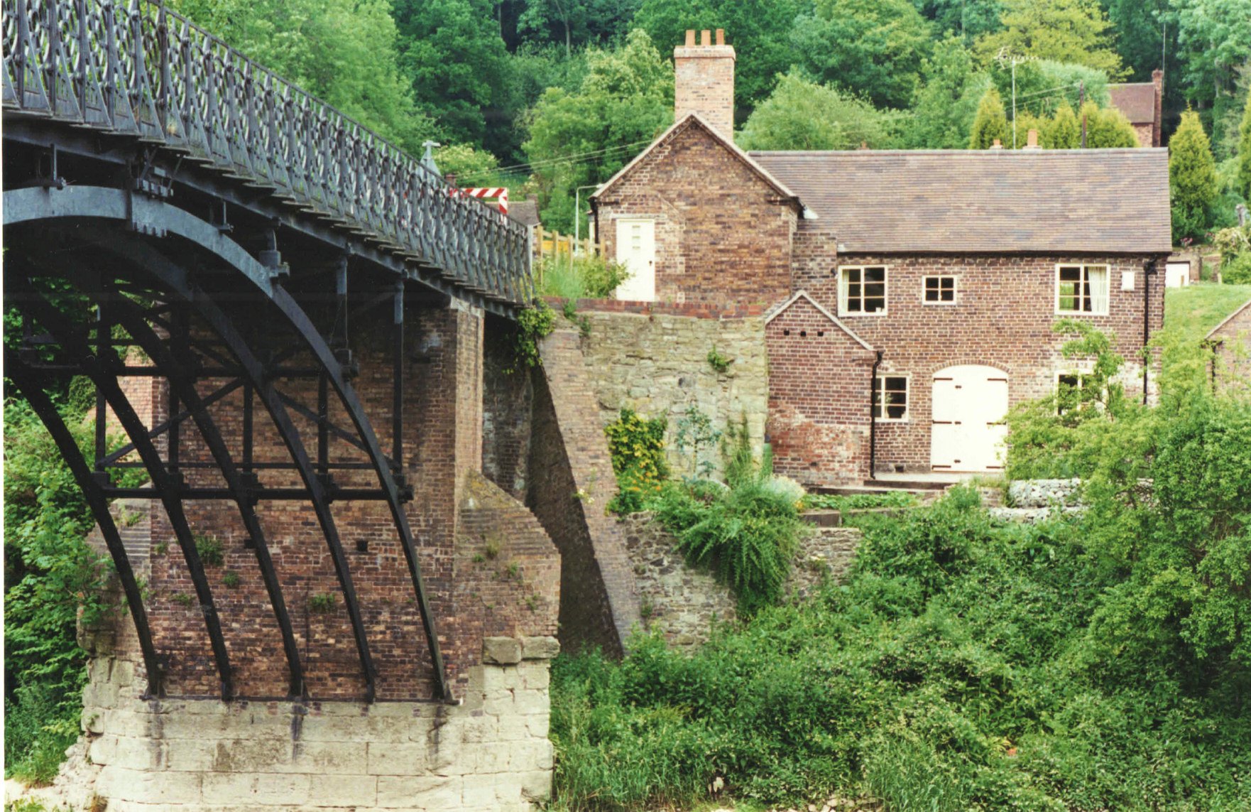 Iron arch and toll cottage are seen together from the Coalport bank in this present-day photograph. Framed like this, it becomes clear how deliberately bridge and gatehouse were designed to function as one — the cottage positioned precisely to let its keeper watch, and control, every crossing.