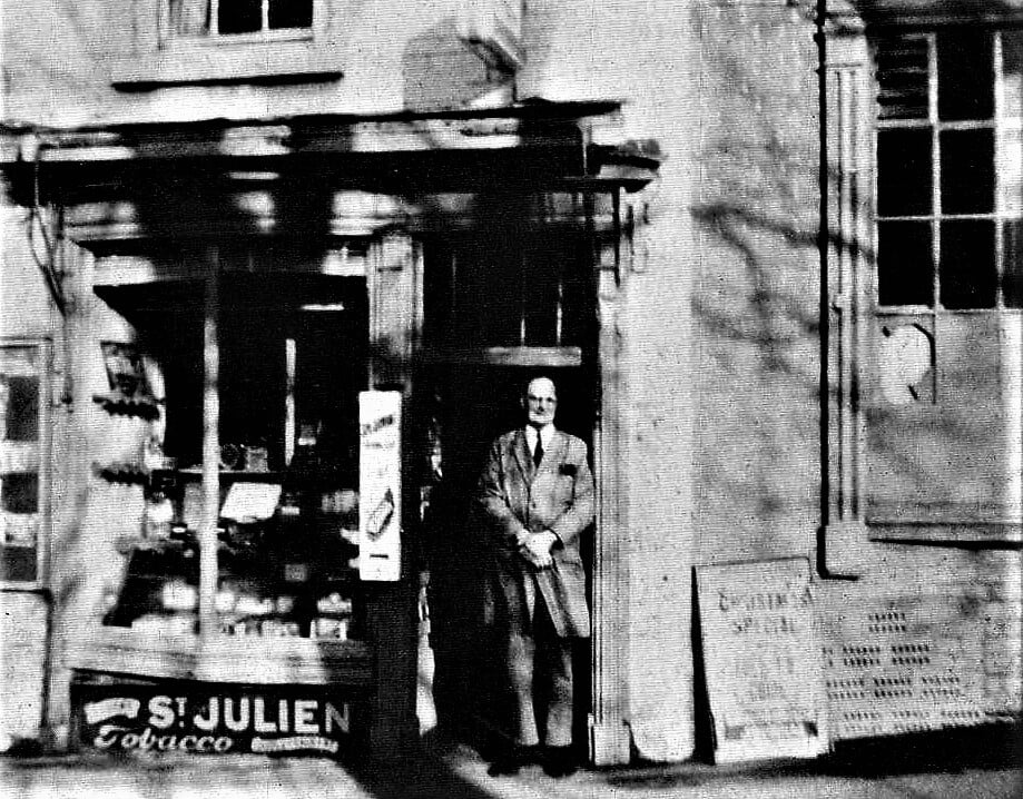 A shopkeeper in his doorway, a St Julien tobacco board propped below the window