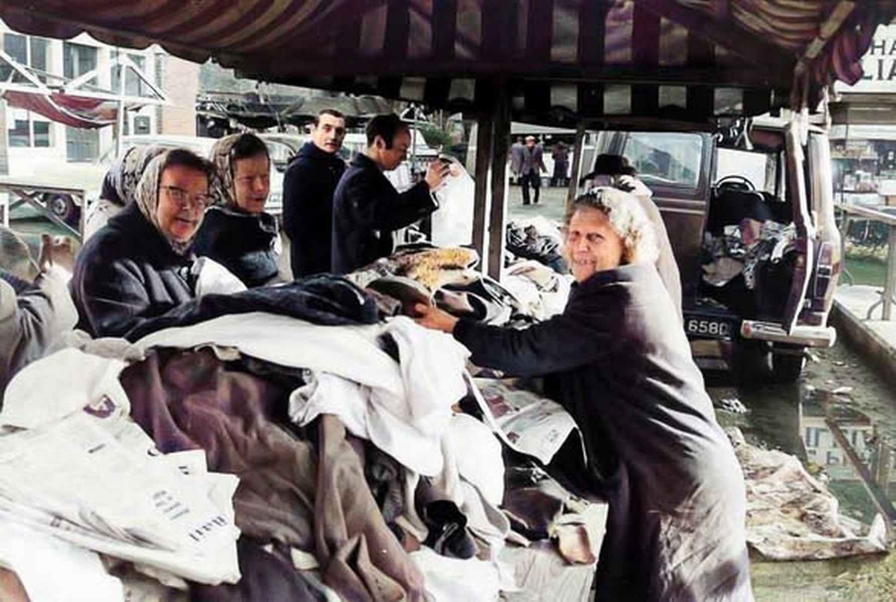 A clothing stall under its awning, garments hung on rails at eye height so shoppers could feel the cloth before buying. Ready-made clothes at market prices were how most families in the Gorge dressed. Exact date unknown, though the styles suggest the middle of the twentieth century.