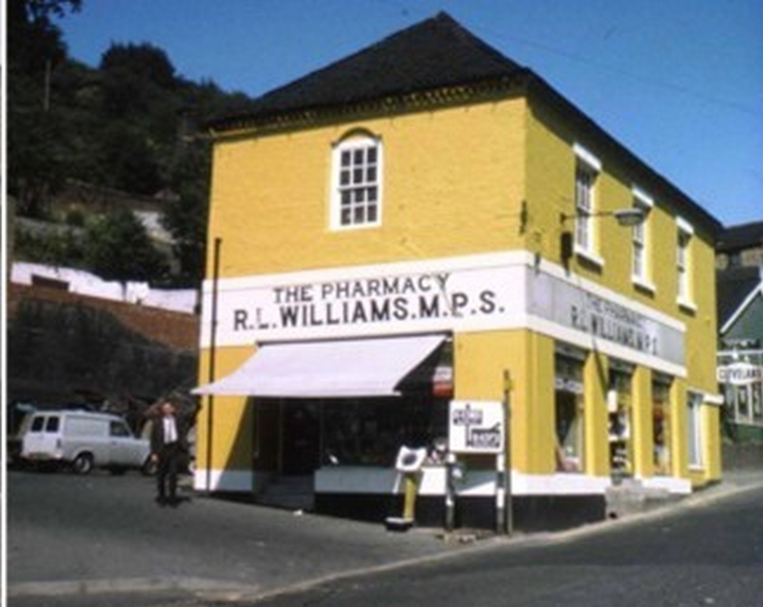 The same shop, now painted yellow and lettered "THE PHARMACY — R. L. WILLIAMS M.P.S.". The parking meter at the kerb is the detail that dates it, and the van beside it agrees: this is Ironbridge in the 1960s or 70s, learning to live with the motor car.