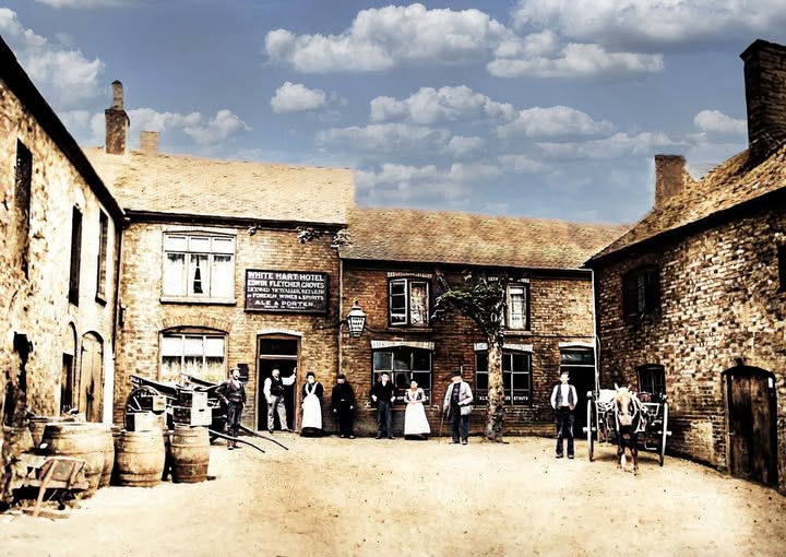 The White Hart Hotel yard, with staff and customers gathered outside, barrels stacked at the left and a horse and cart at the right. Colourised.