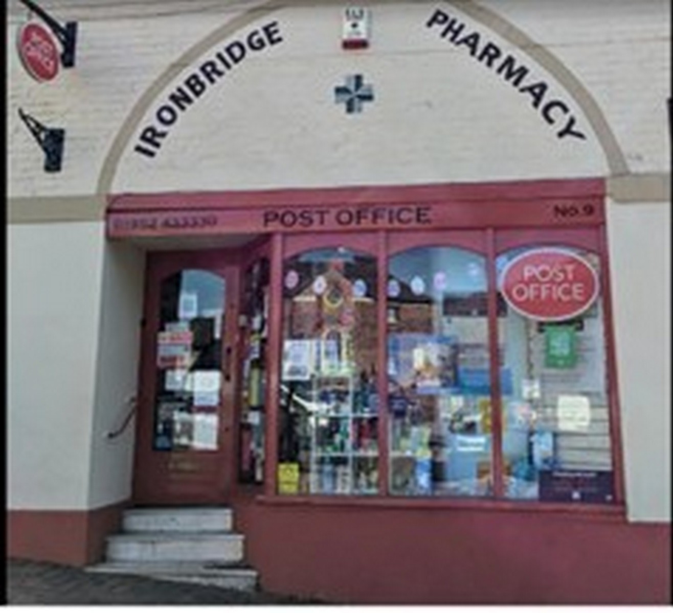 The shopfront tucked under its arch — Ironbridge Pharmacy above, the Post Office below. Two of the services a small town cannot do without, sharing one Victorian opening. The brickwork around the arch is original; only the glass and signage have changed. Present day.